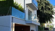2-story modern house with a garage, balcony, and glass elements on the facade. The house has a minimalist, contemporary design with a flat roof and is surrounded by greenery.