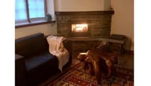 Cozy living room with a stone fireplace, large windows, and a patterned rug on the floor. The room has a comfortable sofa and some decorative elements.