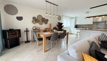 Dining room with a wooden table, gray chairs, a white couch with pillows, a black cabinet, a wine cooler, a kitchen with white cabinets, a countertop, and a hanging lamp.