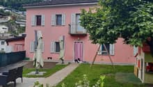 2-story pink house with shuttered windows, balcony, and a small yard with a paved walkway, garden, and outdoor furniture