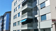 Modern apartment building with multiple balconies, white exterior, blue awnings, and a fenced entrance.