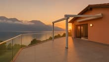 Two-story house with a balcony/terrace overlooking a scenic lake and mountains at sunset. The house has a tiled roof and large windows, with a glass railing on the balcony providing an unobstructed view of the beautiful natural landscape.