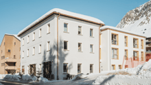 A multi-story white building with a sloped roof, surrounded by snow-covered mountains. The building has several windows and a balcony or terrace visible.