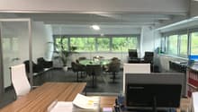 The image shows a modern and spacious office space with large windows providing a view of the surrounding greenery. The office features a glass-enclosed meeting area with a round table and comfortable chairs. There is also a desk with a Lenovo computer an