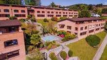 Multi-story brick building with red tile roofs, surrounded by lush gardens, landscaping, and a swimming pool. The building has balconies and terraces overlooking the grounds.