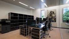 The image shows a modern office space with multiple workstations. The room has a sleek and minimalist design, with black desks, chairs, and storage units. There are several computer monitors and printers on the desks, indicating this is a workspace for of