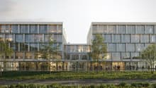 Modern office building with glass facade, multiple floors, entrance with glass doors, people walking around, trees, and green lawn