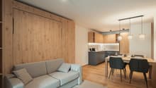 Open-plan living and dining area with a gray sofa, dining table, and wooden flooring. The kitchen is partially visible with a sink and cabinets.