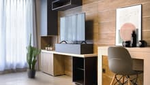 Modern living room with wooden wall panels, a TV mounted on the wall, a desk with a chair, and a potted cactus plant. The room has large windows with curtains, providing a view of a balcony or terrace.