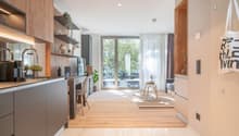 The image shows an open concept living space with a kitchen, dining area, and living room. The kitchen features modern gray cabinets, wooden shelves, and stainless steel appliances. The dining area has a small table and chairs, and the living room has a c