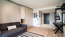 Open-plan studio apartment with hardwood flooring, gray sofa, built-in storage cabinets, and a dining table. The room has a modern and minimalist design with a striped rug and artwork on the wall.