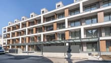 Multi-story apartment building with modern, minimalist design. The building features balconies on each floor, with a mix of glass and wood accents. There is a covered entrance area at the ground level.