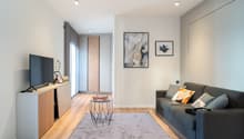 Spacious living room with hardwood floors, white walls, large windows, gray sofa, TV stand, and decorative artwork on the walls. The room also features a sliding door leading to a balcony or terrace.