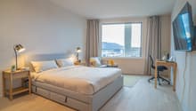 Spacious bedroom with large windows offering a scenic view, hardwood flooring, a king-size bed with storage drawers, two bedside tables with lamps, a desk with a chair, and a flat-screen TV mounted on the wall.