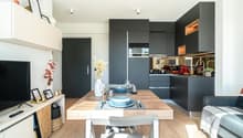 The image shows an open-plan living room and kitchen area with a large wooden dining table, black kitchen cabinets and appliances, and a cozy living space with shelving and decor. The kitchen appears to have a dishwasher visible as one of the appliances.