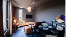 Spacious living room with large windows, brown curtains, wooden TV stand, leather sofa, dining table with chairs, and decorative elements like a fan-shaped wall art piece.