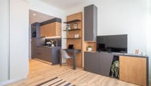The image shows a modern and compact studio apartment with an open-plan layout. The kitchen area features dark gray cabinets and a wooden countertop, with a dishwasher visible. There is a desk area with shelves above it, and a TV stand with storage units.