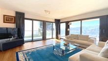 Spacious living room with large windows offering a scenic view, hardwood floors, white sofas, glass coffee table, and a TV stand.