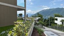Modern building with large windows overlooking a scenic lake and mountains. The building has a balcony or terrace area with lush greenery and landscaping. The surrounding area features a paved walkway and additional landscaping elements.