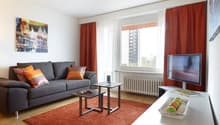 Spacious living room with a large gray sofa, orange accent pillows, and a framed artwork on the wall. The room has a large window with red curtains, providing a view of the city outside. There is a coffee table and a TV stand with a flat-screen TV.