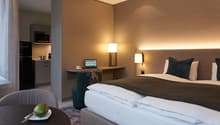 Double bed, bedspread, pillows, lamps, desk, laptop, chair, table, refrigerator, microwave, kettle, kitchen cabinet, grey walls