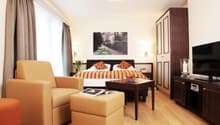 Spacious bedroom with hardwood flooring, large windows, a king-size bed with striped bedding, a seating area with two yellow armchairs and ottomans, a TV stand, and a framed artwork on the wall.