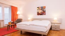 Spacious bedroom with a large bed, wooden floor, orange curtains, side tables with lamps, and a framed artwork on the wall depicting a city scene.