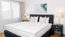 spacious bedroom with large bed, lamps, side tables, nightstands, white linens, framed photo, wooden floor, large window with white curtains