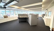 The image shows a modern office space with a large open floor plan. The space has a reception desk, cubicles, and various storage units. The room has large windows that provide natural lighting, and the ceiling has exposed beams and lighting fixtures. The