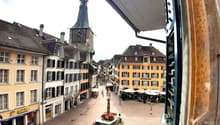 Church clock tower, street with shops, square with fountain, buildings, cobblestone streets, pedestrians