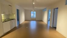 Empty room with white walls, brown floor, kitchen area with sink and cabinets, windows, and doors.
