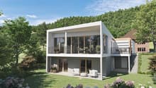 Two-story modern house with a flat roof, large windows, and a balcony on the upper level. The house is surrounded by a well-manicured lawn and lush greenery, including trees and flowering plants. There appears to be a garage attached to the side of the ho