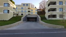 Multi-story apartment building with a garage entrance in the foreground. The building has a modern, minimalist design with a mix of yellow and gray colors. There are balconies and large windows visible on the facade. The surrounding area appears to be a r