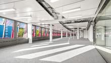 Large open office space with high ceilings, exposed ductwork, and floor-to-ceiling windows providing natural light. The space has a modern, minimalist design with grey carpeting and white walls. There are multiple columns throughout the space.