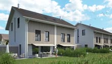 Two-story modern house with gray siding, large windows, and a balcony. The house is surrounded by a grassy yard and has a paved parking area.