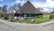 A traditional German-style half-timbered house with a large pyramidal roof, surrounded by trees and a grassy area. There is a parked car in the driveway in front of the building.