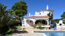 Two-story Mediterranean-style villa with a red tile roof, white exterior walls, and a tower feature. The house is surrounded by lush greenery, including palm trees and other vegetation. There is a swimming pool in the foreground with a patio and outdoor s