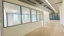 The image shows a large open office space with white walls, windows, and a concrete floor. The space appears to be unfinished, with exposed ceiling beams and lighting fixtures. The room has multiple glass-paneled walls, creating separate sections or offic