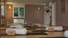 A yoga studio with yoga mats, bolsters, pillows, and lamps on the ceiling. The floor is wooden, and the walls are painted in a muted purple. There is a shelf with towels, a potted plant, and a framed artwork.