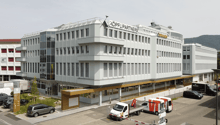 white exterior, multiple windows, 'Koepfli Partners' sign, cars parked in front, adjacent buildings, mountains in the background