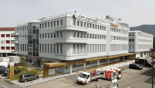 white exterior, multiple windows, 'Koepfli Partners' sign, cars parked in front, adjacent buildings, mountains in the background