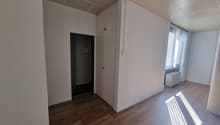 The image shows a simple, minimalist room with white walls, a suspended ceiling with a grid pattern, and a wooden floor. The room has a doorway leading to another area. The room appears to be empty, with no furniture or decor visible.