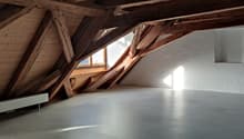 Spacious attic with exposed wooden beams and trusses, large windows providing natural light, concrete floor, and potential for conversion or storage space.