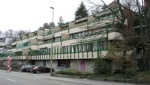 Multi-story apartment building with balconies, brick and concrete construction, surrounded by trees and vegetation