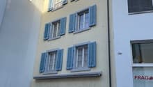 An apartment building with several windows and blue shutters, a yellow painted wall, a black door at the entrance and a display glass in the bottom floor.
