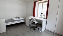 room features a single bed, desk with a chair, a window with red curtains, and a lamp. White walls and wooden floors.