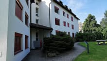 White multistory building with red windows, brick pathway, green grass, bush, bench