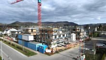 Aerial view of a construction site with multiple buildings under construction, a large red crane, scaffolding, and a road with parked cars.