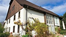 two-story house, brown roof, white wall, several windows, multiple potted plants, white lawn umbrella, greenery