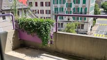 Empty balcony with concrete floor and railing, overlooking a street with trees, houses and hills.